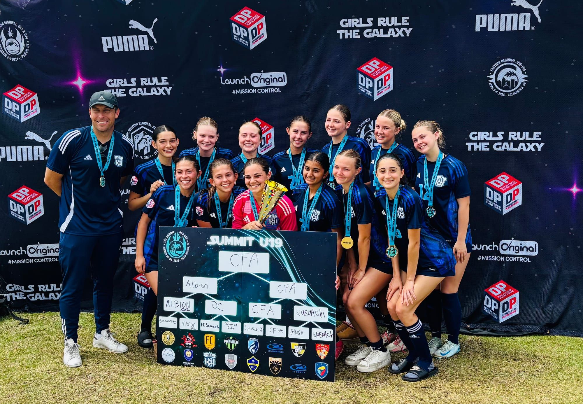 CFA girls team with medals and trophy in front of a tournament banner